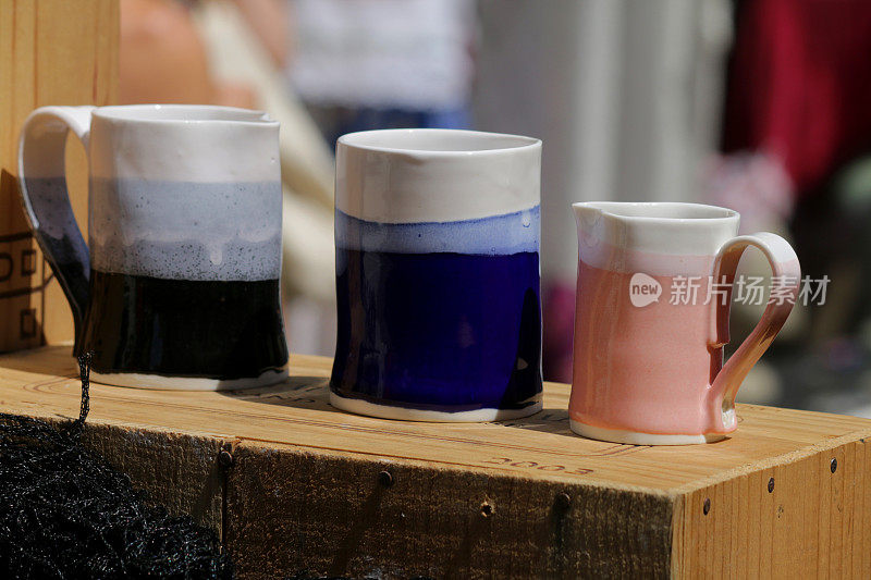 木盒上放着三排自制/手工陶制的陶杯和陶罐，放在陶制轮上，上釉，然后在窑中烧制，作为咖啡/茶的马克杯组合，杯子的颜色有深蓝色、天蓝色和粉色